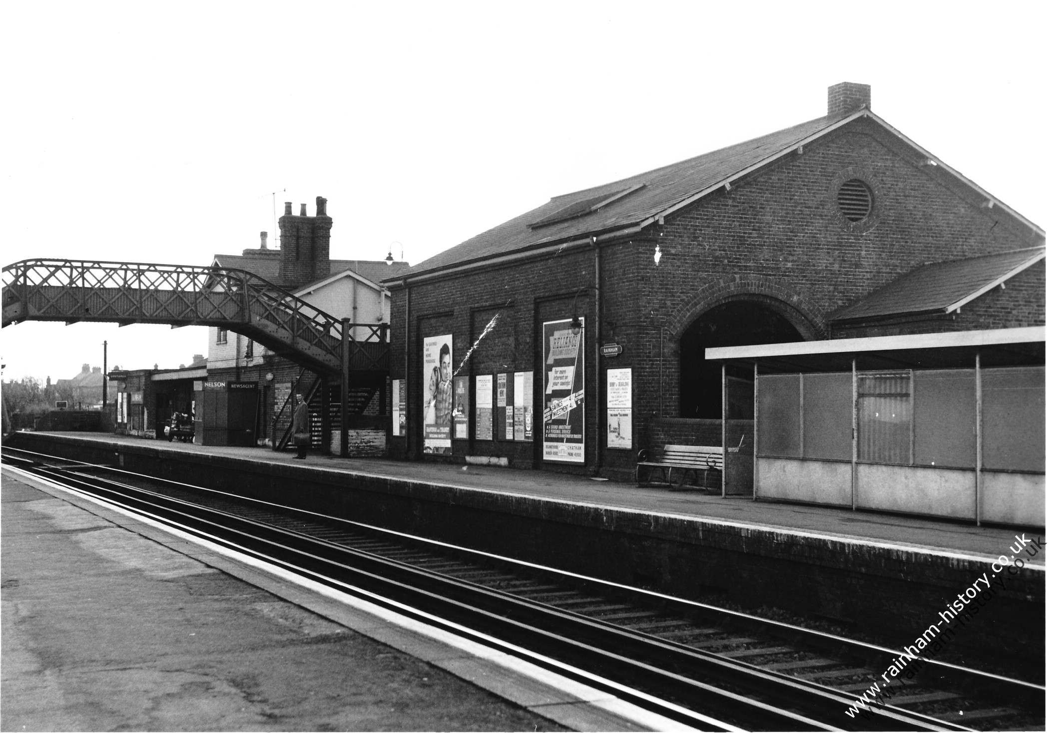 view of the Station buildings from the coastbound platform with the footbridge over the tracks and a passenger waiting on the platform. There is a poster for Chatham Reliance Building Society.